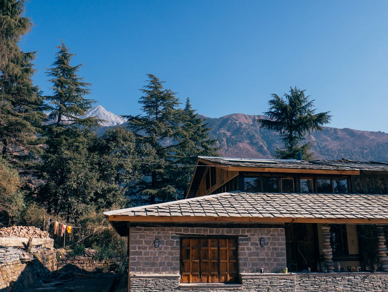 An old stone house with a grey slate roof and wooden upper floor among deodar trees, with mountains behind under a clear blue sky.