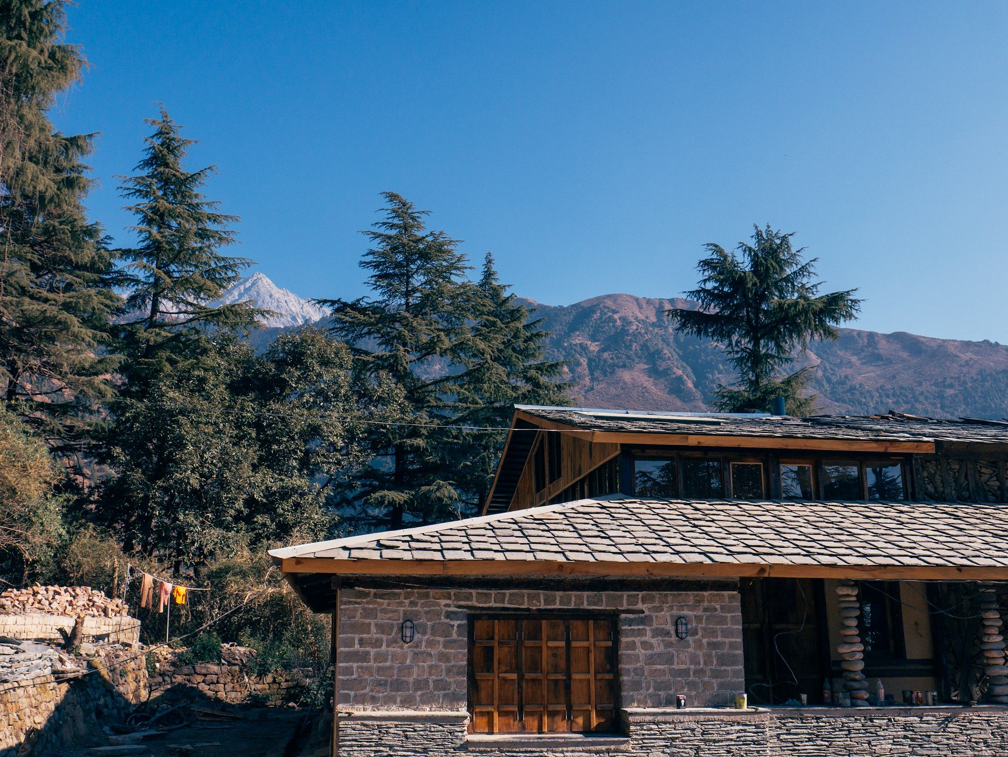 An old stone house with a grey slate roof and wooden upper floor among deodar trees, with mountains behind under a clear blue sky.