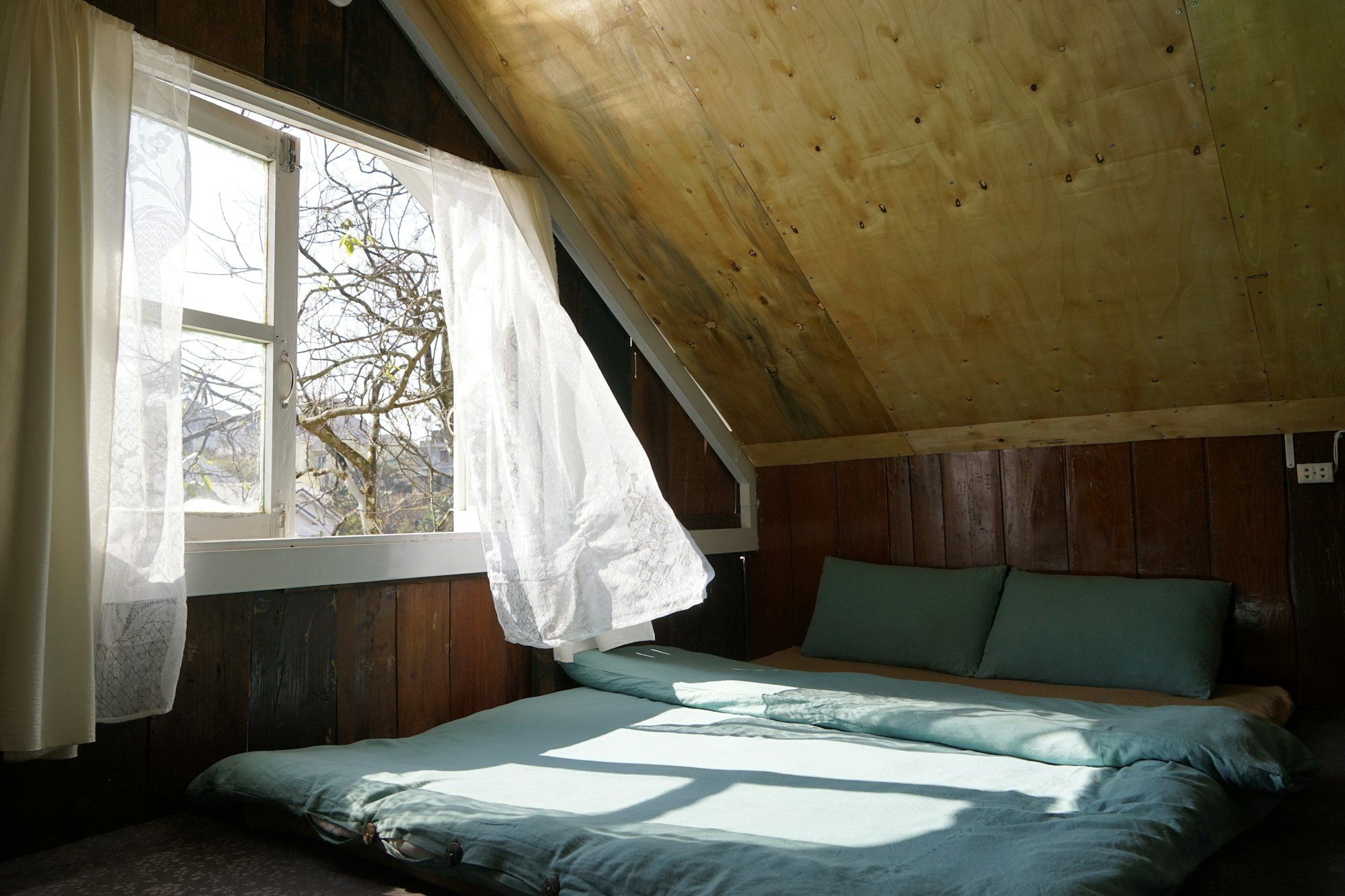 A low bed under a sloping wooden ceiling, with light coming through a window and a white curtain.