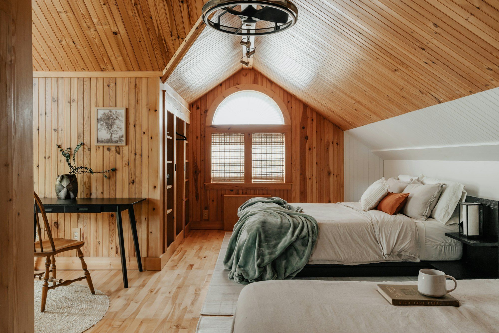 A room lined in pale wood under a pitched ceiling, with two beds, a wool throw and an arched window.