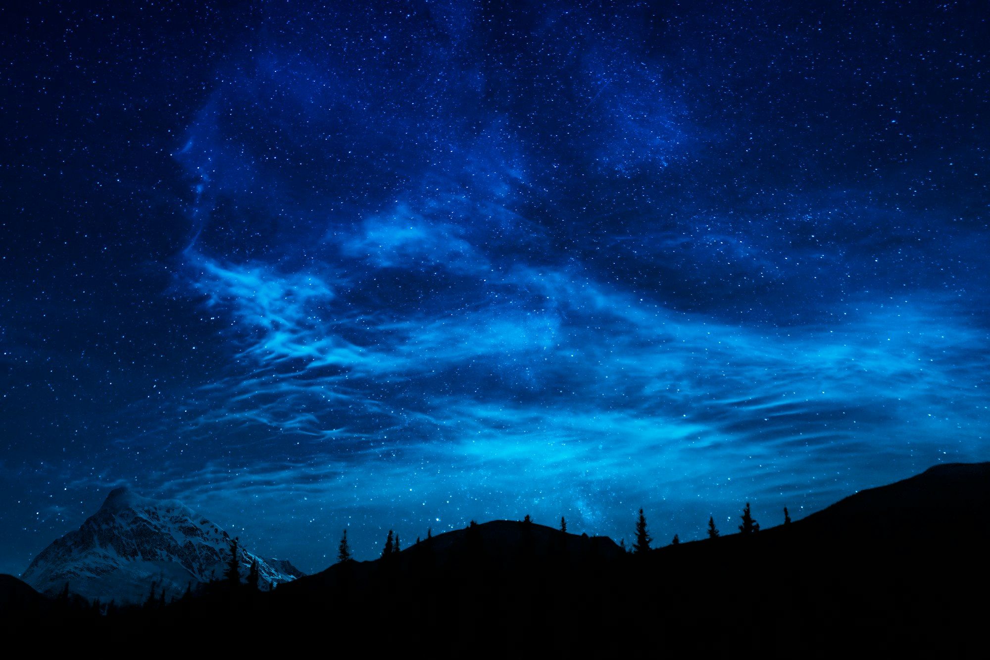A night sky full of stars above the dark outline of trees and hills.